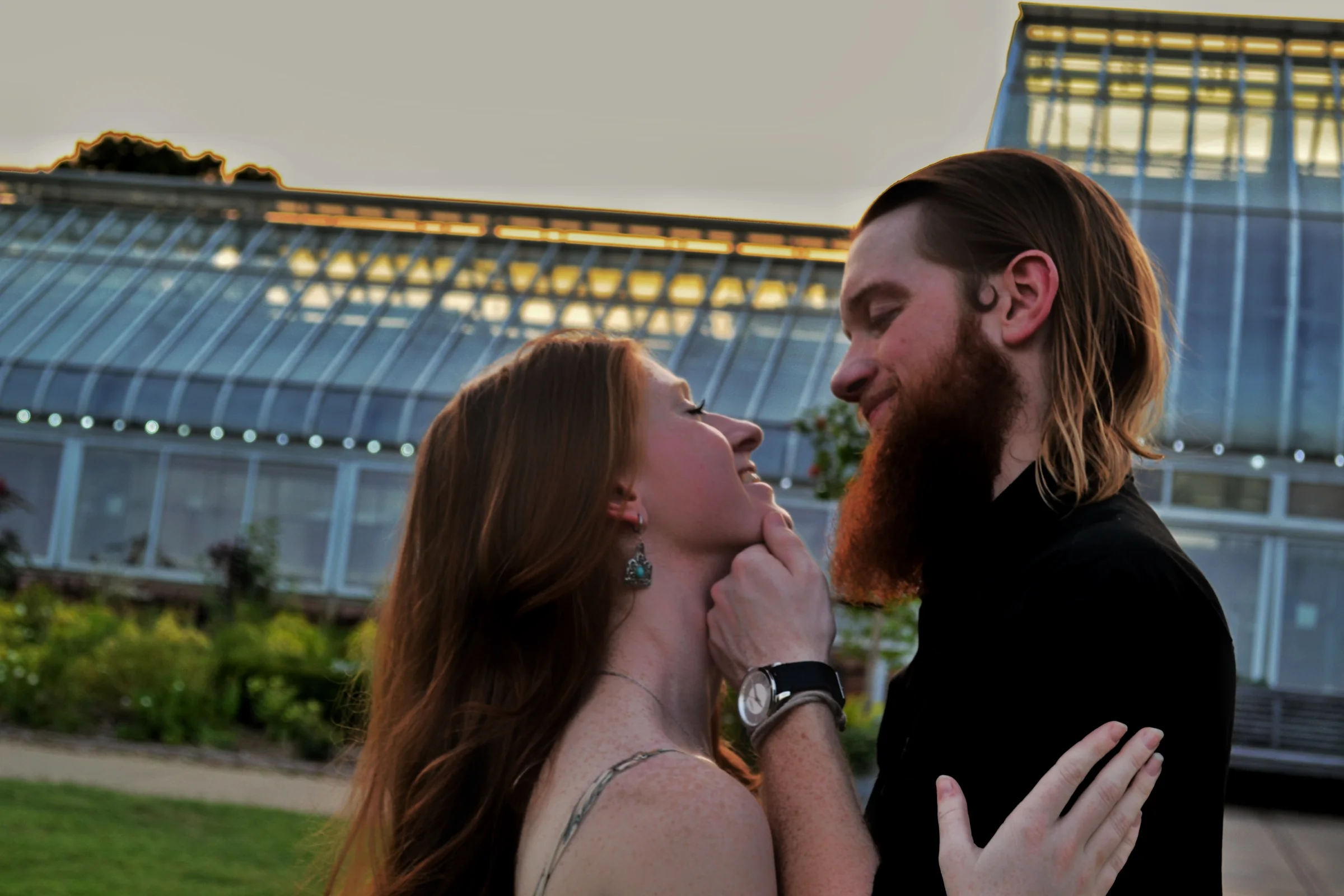 A couple smiling closely at one another outside a greenhouse at dusk.