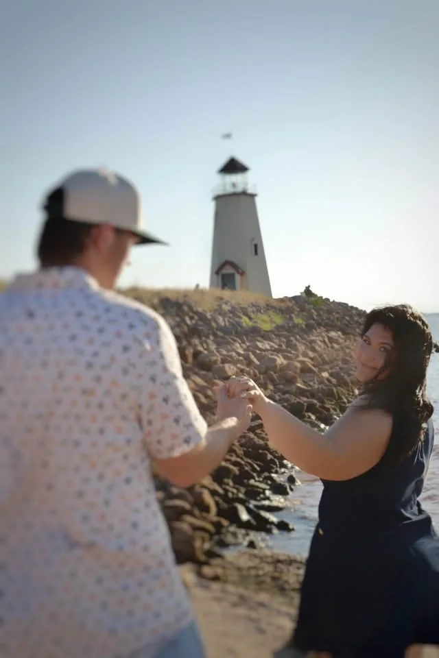 A couple holding hands on a rocky shoreline beneath a lighthouse.